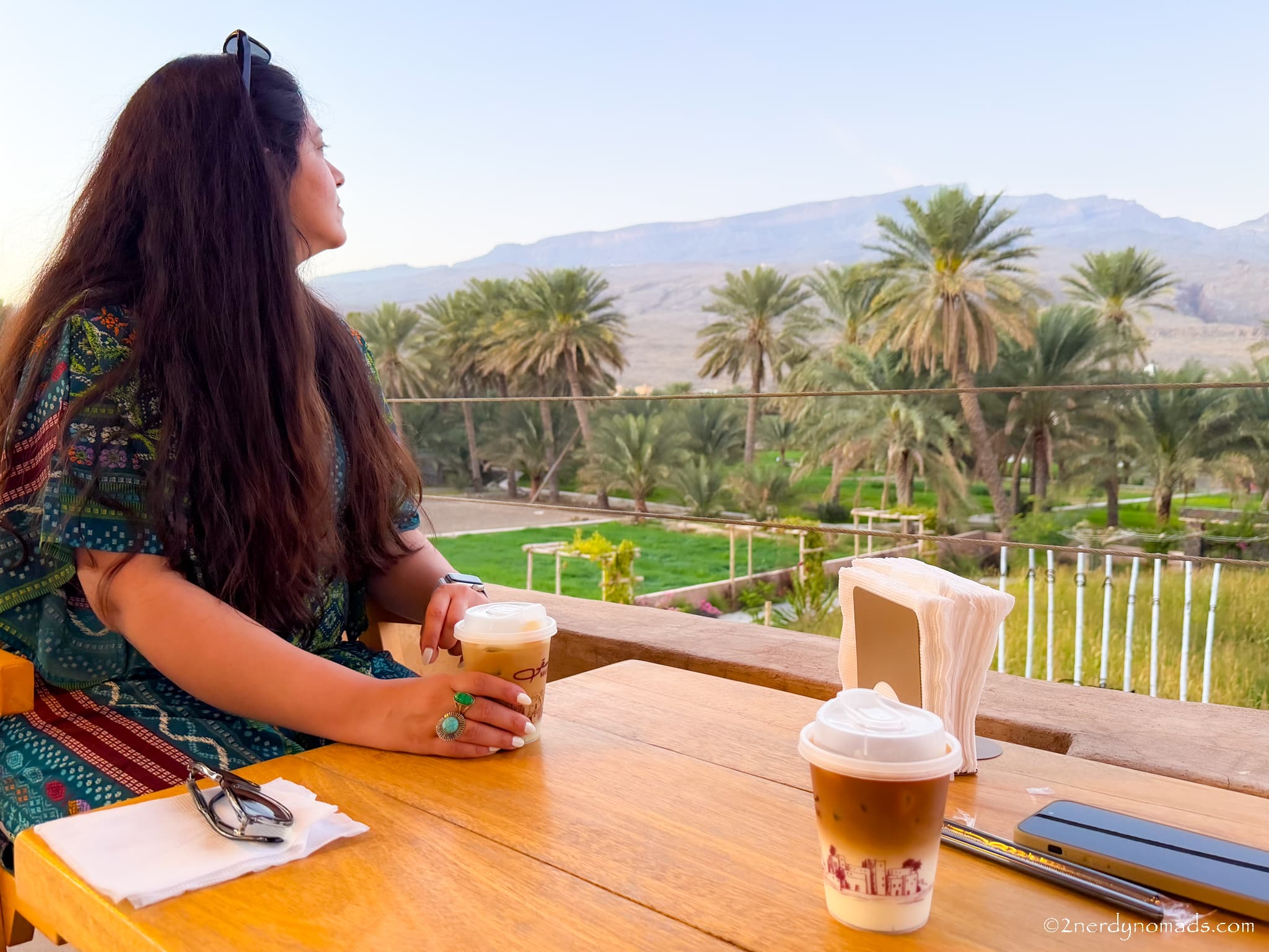 Sitting at a café in Al Hamra, Oman, overlooking the Hajar Mountains and lush palm trees.