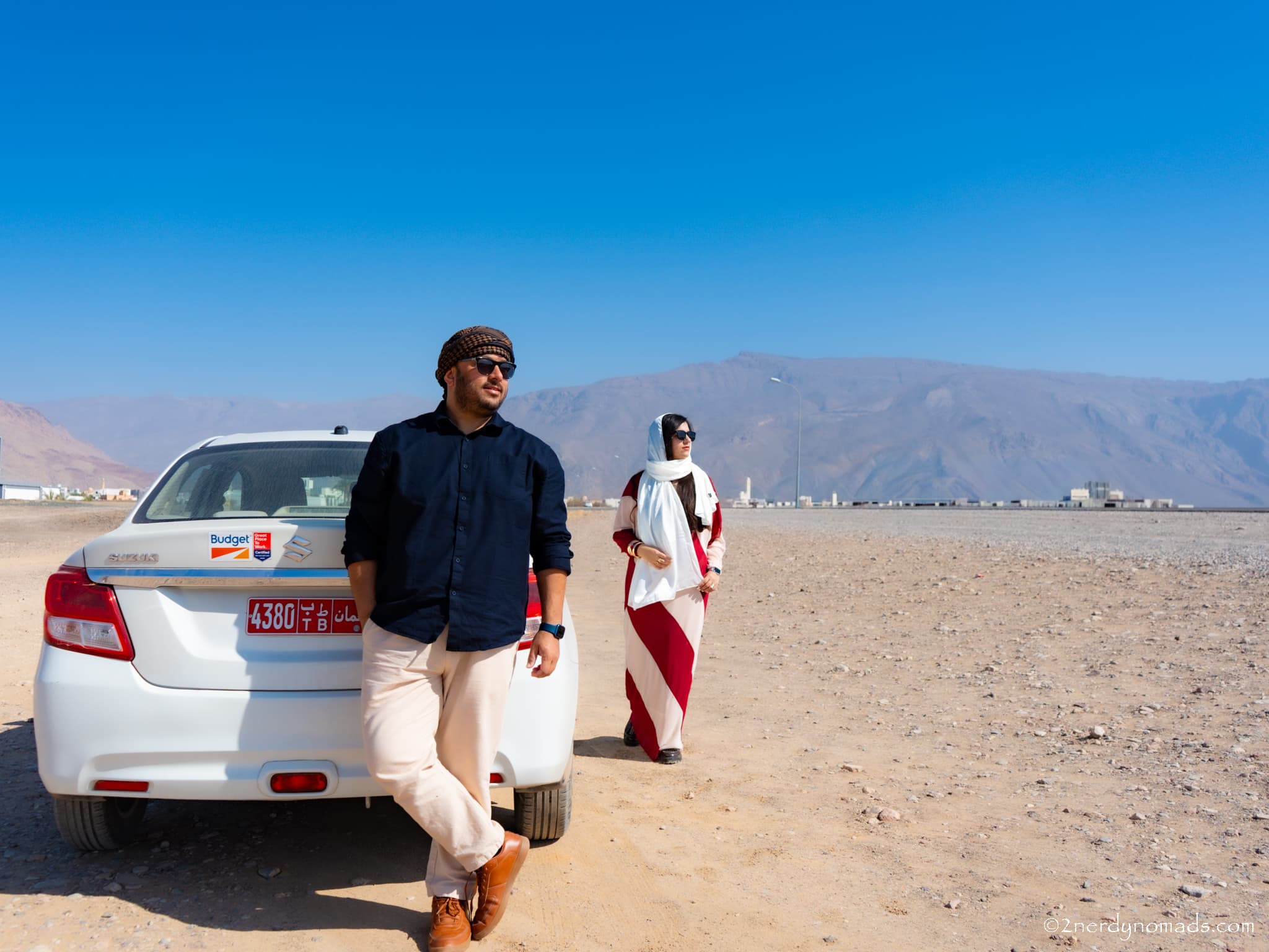 Urvi and Animesh standing in front of their car with the rugged mountains of Oman in the background.